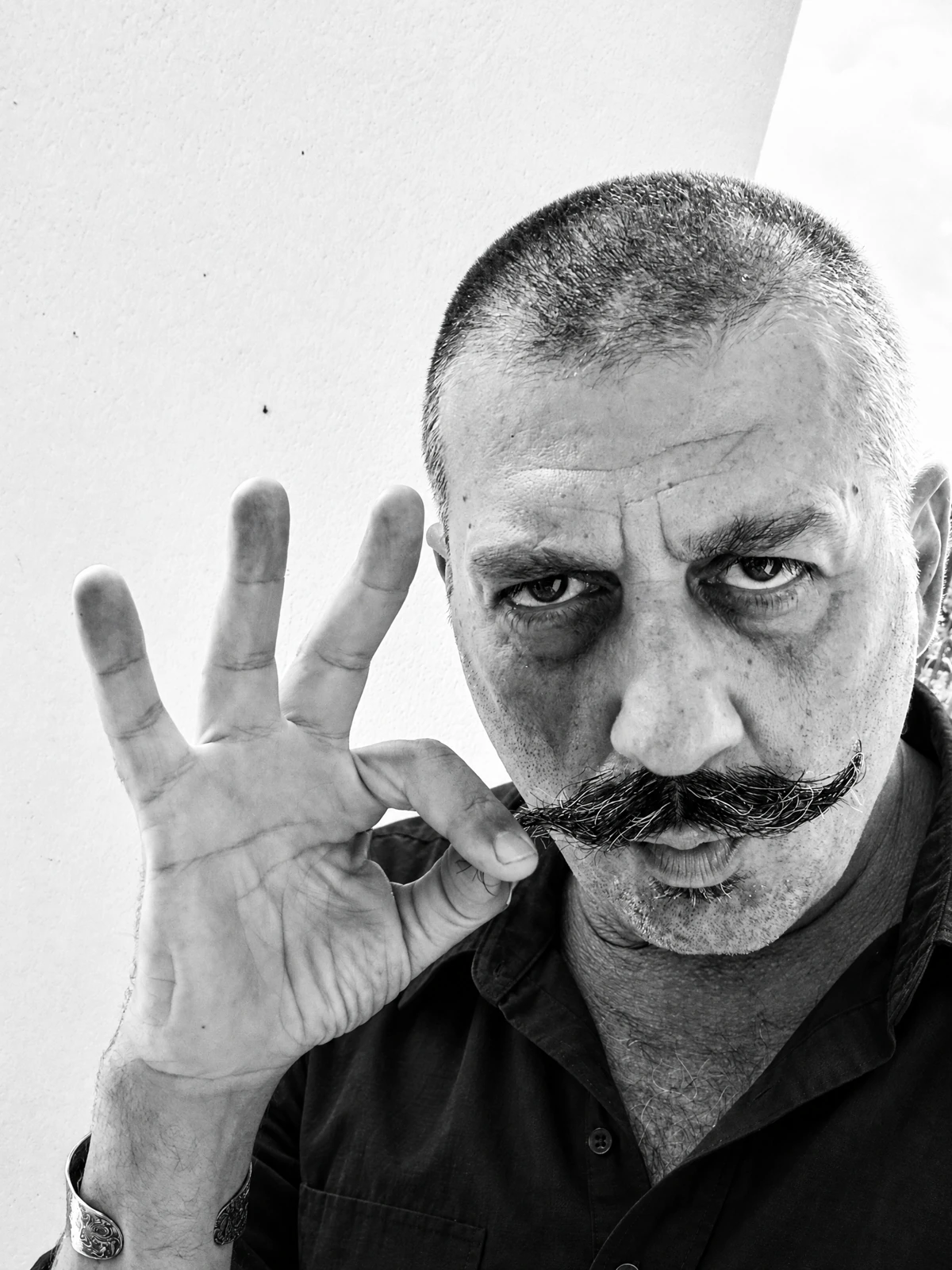 Black-and-white portrait of Oz Fikri in a plain black shirt, with his distinctive moustache and an OK hand gesture